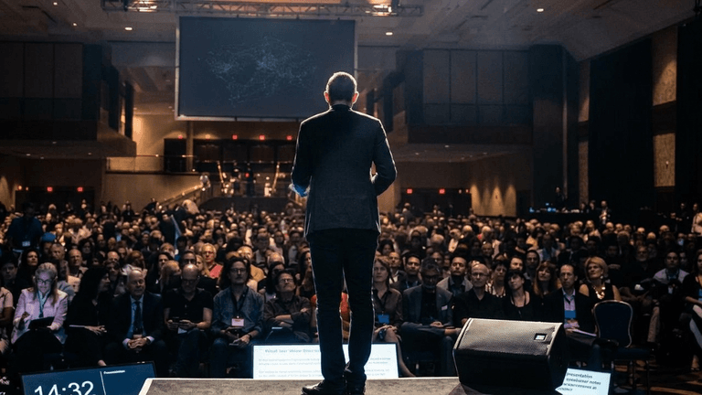 Speaker at a conference addressing a large audience in a dimly lit venue