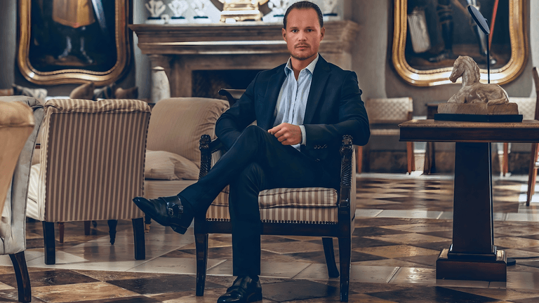 A confident man in a tailored dark suit sits cross-legged in an elegant, classical lounge with marble floors and antique décor.