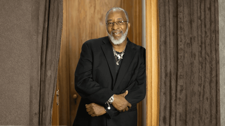 Smiling man with gray beard and glasses in black suit leaning in doorway between curtains.