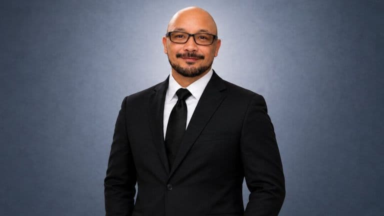 Professional portrait of Charles Santos wearing a black suit and tie against a gray background.