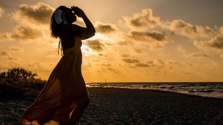 A woman with a flower in her hair stands on a beach at sunset, embodying peaceful reflection and global perspective.