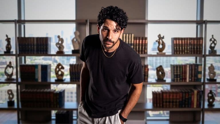 Man with curly hair, wearing a black t-shirt, standing in front of a bookshelf with a city view.