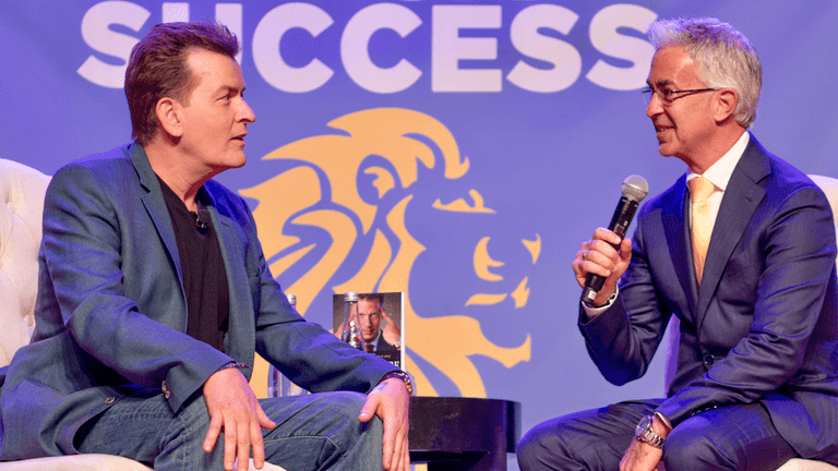 Craig Carr speaks on stage with another man during a business success event promoting psychology-based wealth and sales strategies.
