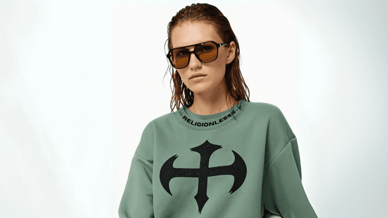 "Model wearing a green sweatshirt with the Religionless logo on the front and bold sunglasses, posing confidently against a simple white background."