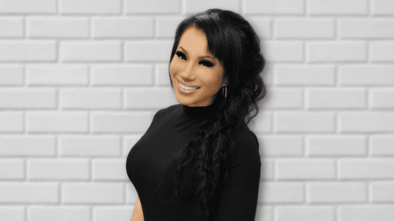 A smiling woman with long, curly black hair stands in front of a white brick wall, wearing a black long-sleeve top and gold hoop earrings, radiating confidence and positivity.