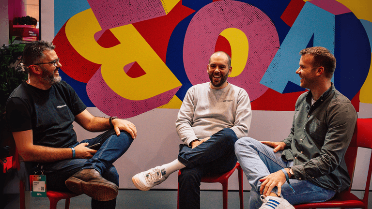 Three men sitting and laughing together in front of a colorful background at a business event, discussing ideas.