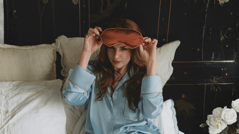 A woman in light blue silk pajamas holds a sleep mask, sitting on a bed surrounded by soft pillows and flowers.