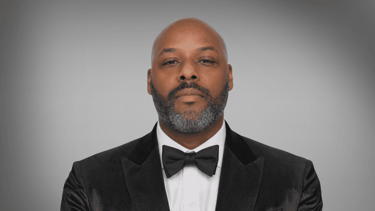 Man in black tuxedo and bow tie against gray background, formal portrait.