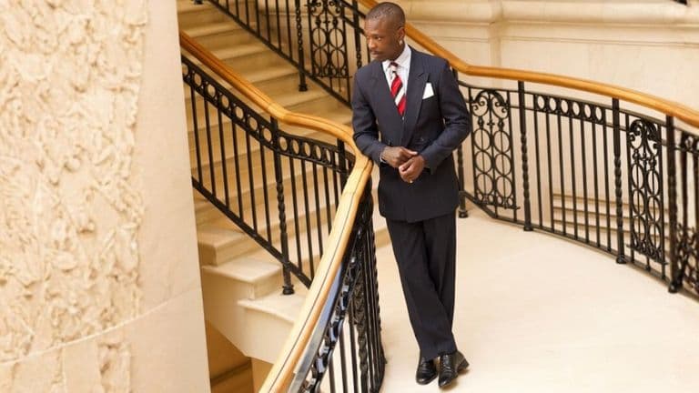 A man in a dark suit, white shirt, and red striped tie stands on a grand staircase with ornate railings, exuding confidence and elegance.