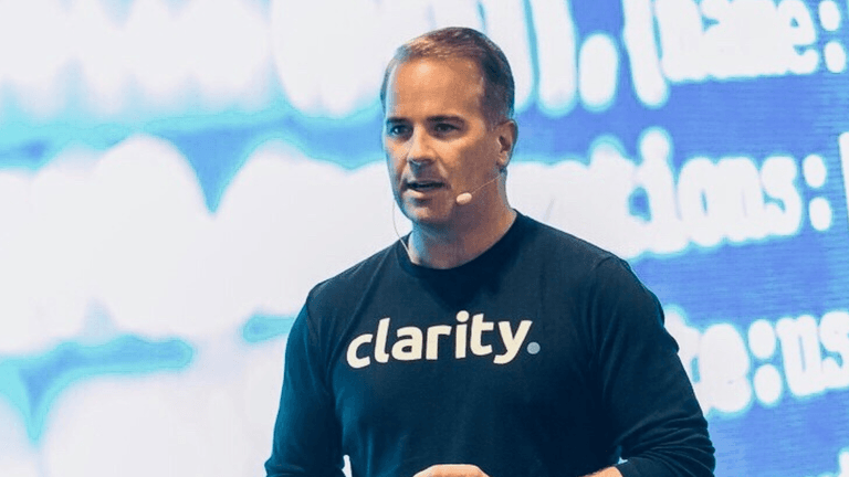 Man speaking at a conference wearing a 'clarity' branded shirt, with a digital background.