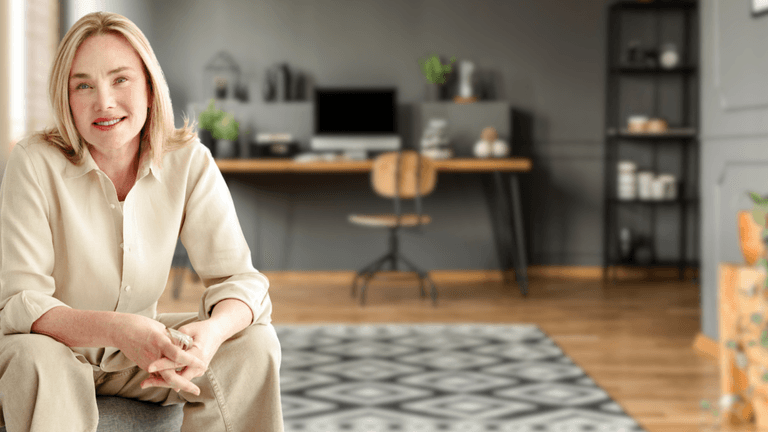 "Smiling woman in a beige blouse and pants, sitting comfortably on a chair in a modern, stylish home office with a wooden desk, computer, and plants in the background."
