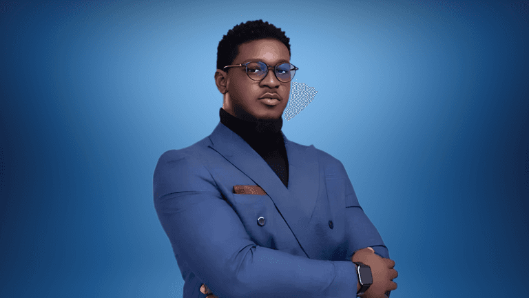 "Professional portrait of a man in a blue suit, wearing glasses, with his arms crossed confidently. The background features a gradient blue tone, creating a sleek and modern look."