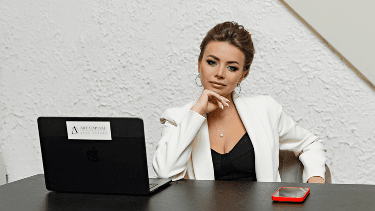 Olga Alekseeva, founder of Art Capital Real Estate, sitting at a desk, reflecting on real estate strategy, with laptop and phone.