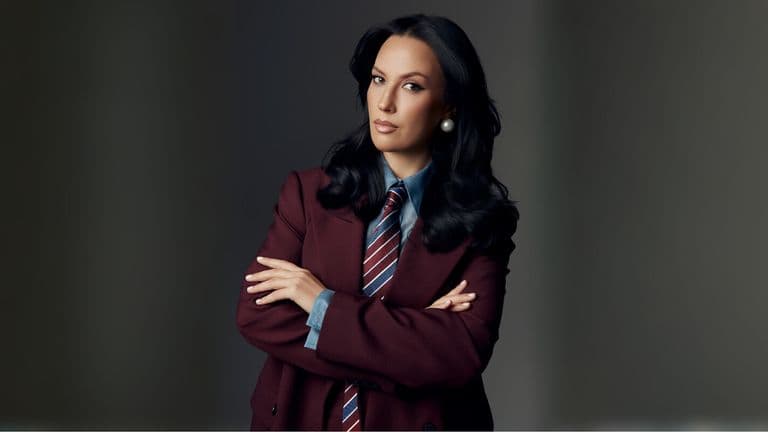 Confident woman in a tailored burgundy suit with a striped tie, exuding professionalism and leadership, arms crossed.