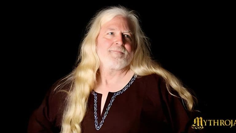 A man with long white hair and a beard, Keith Wescovich, stands in a medieval workshop surrounded by crafting tools.