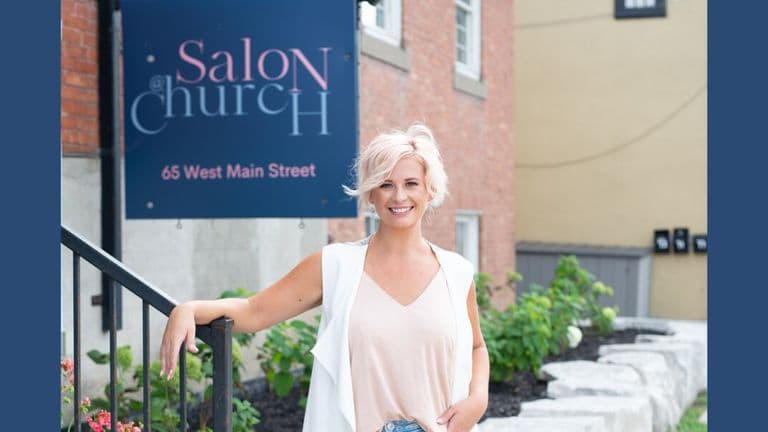 A smiling woman with short blonde hair stands outside a building with a sign reading "Salon@Church" on Main Street.
