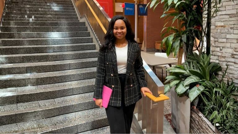 A professional woman in a checkered blazer and black pants stands confidently by a staircase, holding a pink tablet.