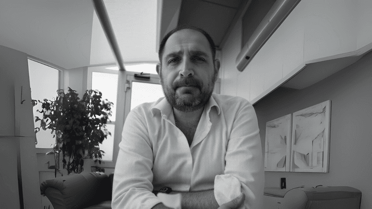 "Professional portrait of Emiliano Caradonna, wearing a white shirt, sitting with arms crossed in a modern office setting with a plant and minimalistic decor in the background."