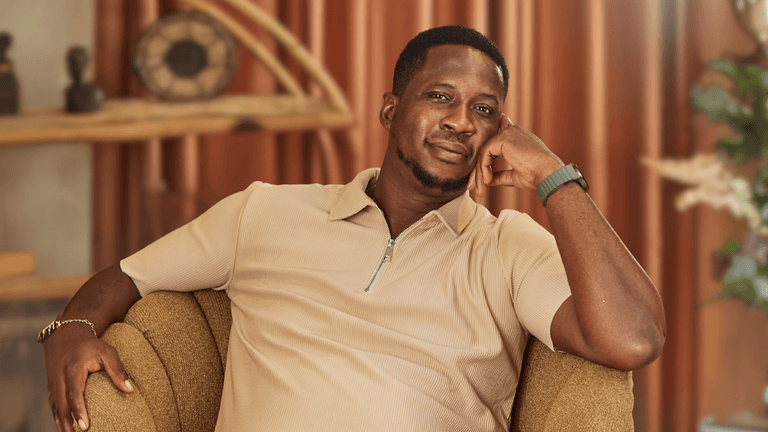 "A confident man in a beige polo shirt smiles while resting his head on his hand, seated comfortably on a stylish armchair in a modern, cozy interior with soft lighting and decorative plants."
