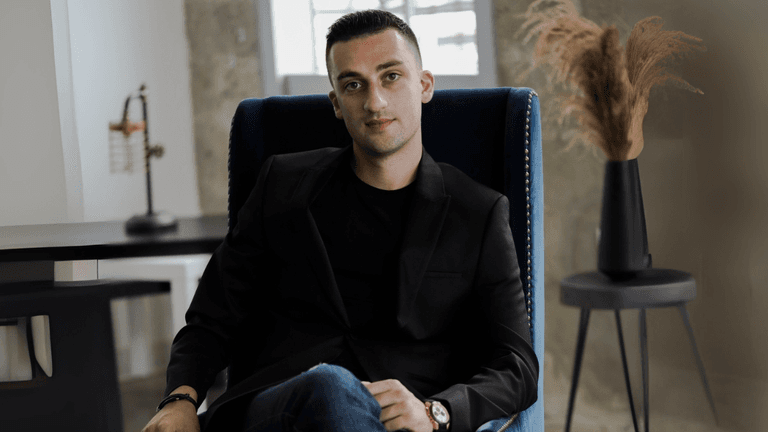 A man in a black blazer and jeans sitting confidently in a blue chair with a minimalist office background.