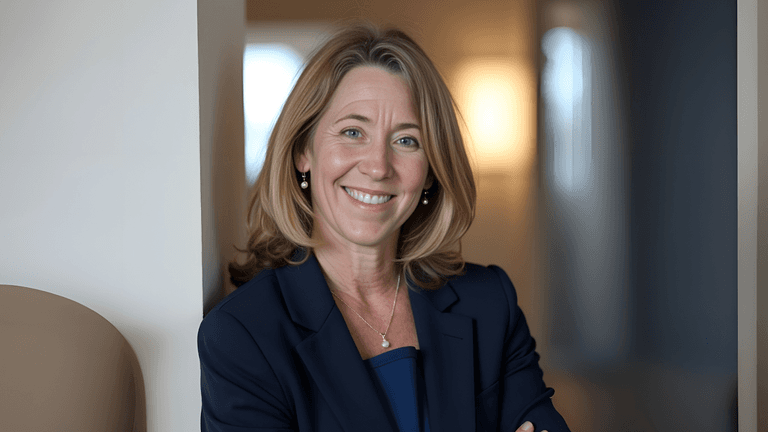A smiling woman in a professional navy blazer stands with arms crossed in a modern office environment.