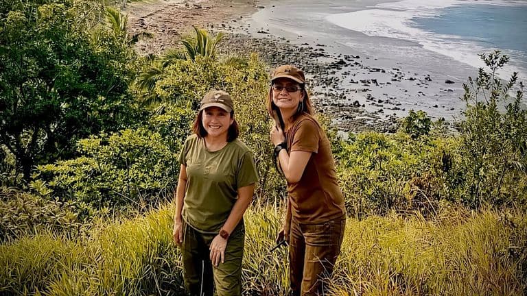 Two women hiking through lush greenery with a scenic beach view in the background.