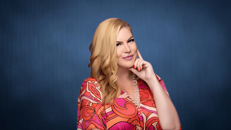 Professional portrait of a woman in a colorful patterned blouse, smiling and resting her chin on her hand with a confident expression against a blue background.