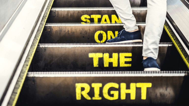 Person walking up an escalator with the words 'STAND ON THE RIGHT' clearly visible in yellow on the steps.