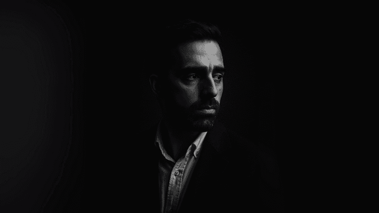 Black and white portrait of a man in a dark suit, looking thoughtfully to the side, with dramatic lighting emphasizing the contours of his face and creating a moody, contemplative atmosphere.