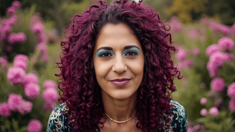 A woman with curly purple hair and blue eyeshadow, smiling in front of a background of pink flowers.