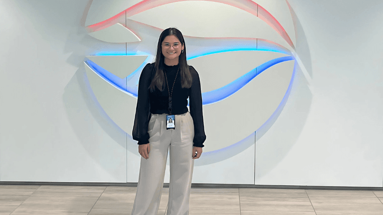 Professional young woman smiling confidently in front of a modern, illuminated company logo, dressed in business attire with light-colored trousers and black top."