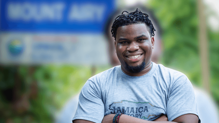 Smiling man wearing a “Jamaica Story” shirt stands in front of a Mount Airy sign.