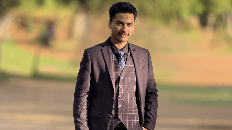 "Young man in a brown suit with a plaid pattern, standing confidently in a park with blurred greenery in the background, showcasing a professional appearance."