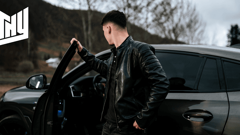 "Young man in a black leather jacket standing next to a car, opening the door while looking away, with a scenic background of hills and trees."