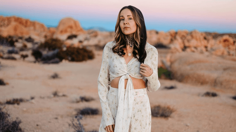 A woman stands in a desert landscape, wearing a white flowy outfit, with a calm expression