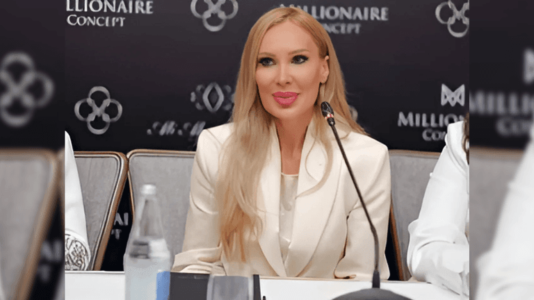 Laura Svag speaking at a press event, wearing a white suit with a microphone in front of her.