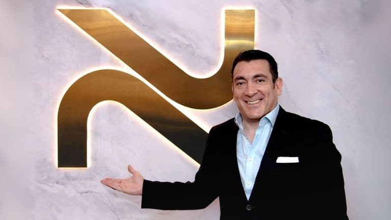 A man standing next to a glowing NovoSculpt logo on a marble wall, smiling and presenting the brand with a welcoming gesture. The logo represents the NovoSculpt fat removal technology.