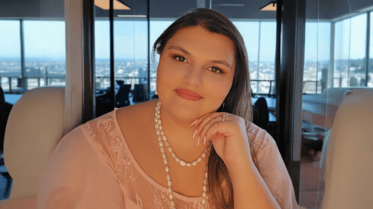 A woman with long hair and pearl jewelry, sitting and smiling while posing with her hand on her chin. She is in a modern office with a panoramic view of the city in the background.