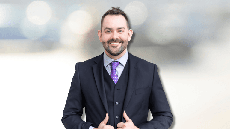 Professional portrait of a man in a suit with a purple tie and a confident smile. He stands with his thumbs up, set against a blurred background of soft lights, portraying a confident and approachable business image.