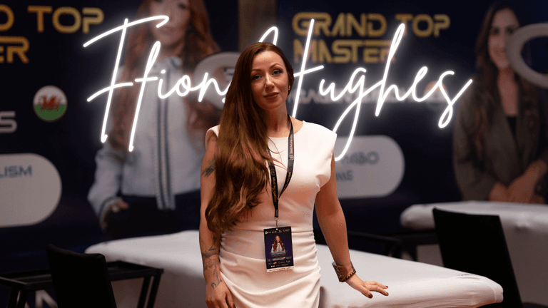 Ffion Hughes, a CEO and entrepreneur, standing confidently at an event. She is wearing a white dress and has long hair. With a badge around her neck, she poses in front of a promotional banner with her name, "Ffion Hughes," illuminated in neon lights. The background suggests a professional event related to entrepreneurship and beauty, reflecting her leadership in empowering women in business.