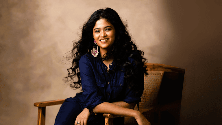 Smiling woman with curly hair in a navy blue dress and statement earrings, sitting on a chair in a relaxed pose, ideal for content related to lifestyle, beauty, and personal empowerment.