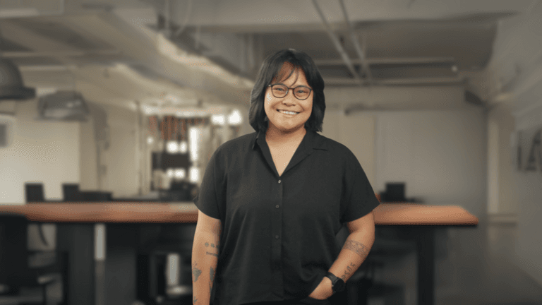Portrait of a woman with short black hair and glasses, smiling confidently in a black button-up shirt, standing in an office setting with blurred background.