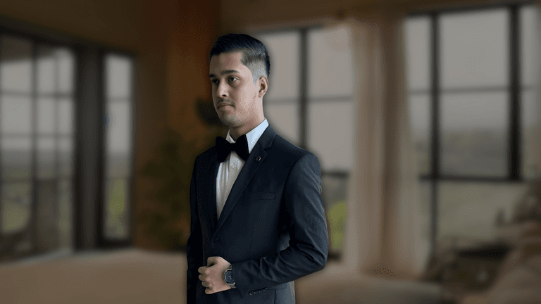 Man in formal black tuxedo and bow tie standing in a well-lit modern living room with large windows, showcasing a sophisticated look.