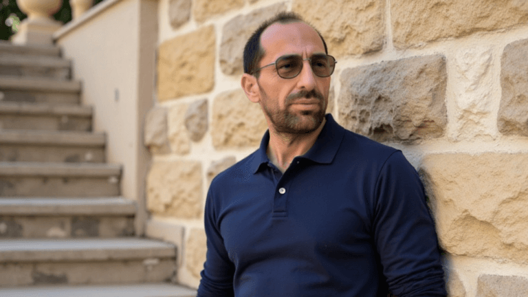 Man in navy shirt and sunglasses standing beside stone wall near stairs.
