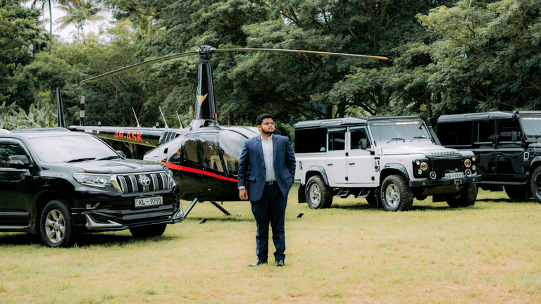 Man in a blue suit standing in front of a helicopter and a lineup of black and white off-road vehicles, set in a scenic outdoor location.