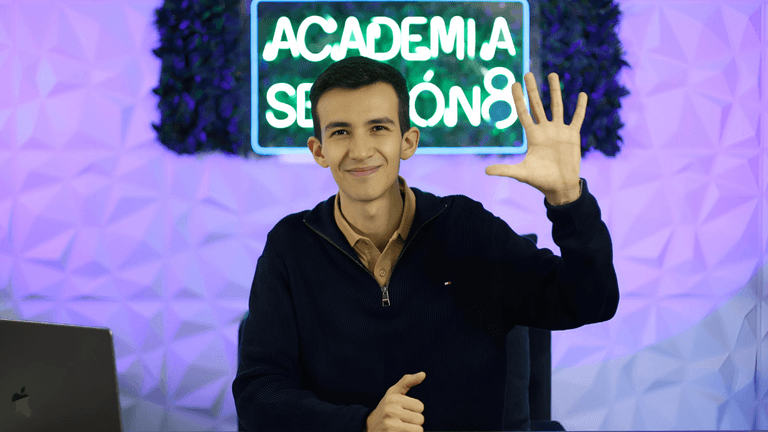 Smiling young man wearing a navy sweater and beige shirt, sitting at a desk with a MacBook in front of him. He is raising his right hand with five fingers up, posing in front of a neon sign that reads 'Academia Secreta' with a modern geometric background.