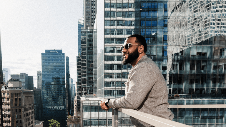 A man standing on a rooftop terrace with a cityscape background, smiling while wearing sunglasses, with modern glass buildings reflecting the sunlight.