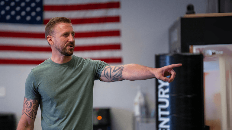 Man with tattoos pointing while speaking, with American flag in background.