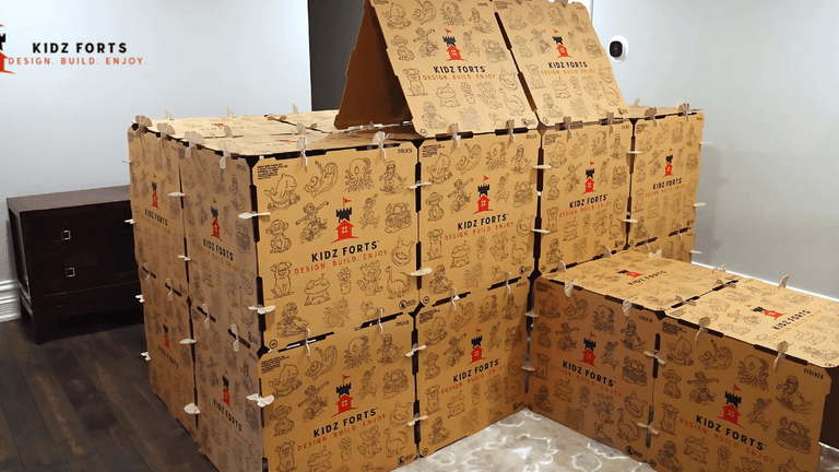 A large, creative fort built from Kidz Forts cardboard building kits, featuring multiple stacked boxes with playful designs and illustrations. The fort has a pitched roof and is designed for children to build and enjoy.
