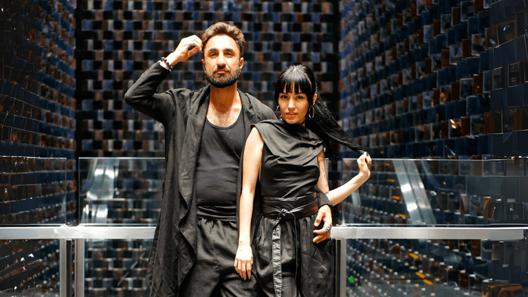 A stylish couple posing in black outfits against a backdrop of mirrored walls with a modern, geometric design.